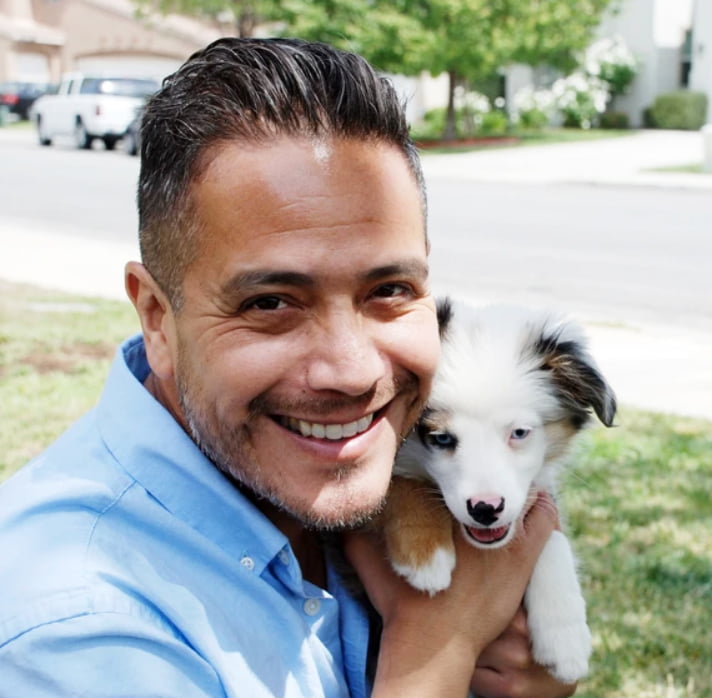 A man in a blue shirt smiles while holding a small white and brown puppy outdoors on a sunny day, capturing the warmth of home.