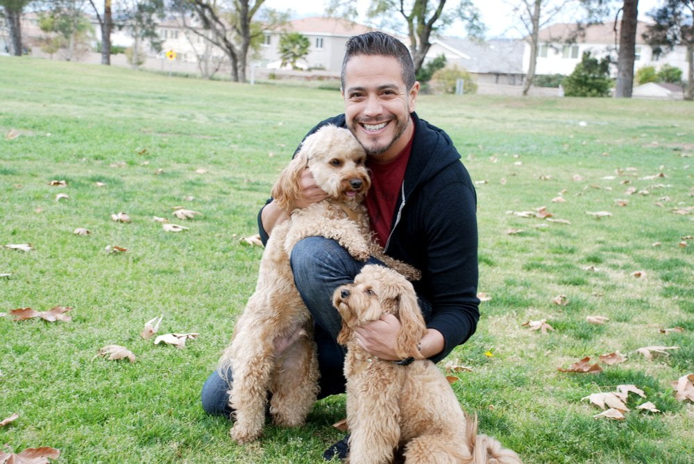 A man crouches on a grassy field, smiling and holding two light brown fluffy dogs. Trees and houses are visible in the background, giving a sense of home.