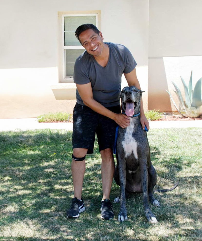 Man in a grey t-shirt and black shorts smiling and kneeling beside a large grey Great Dane in a yard with a house in the background. For more heartwarming moments, contact us today!