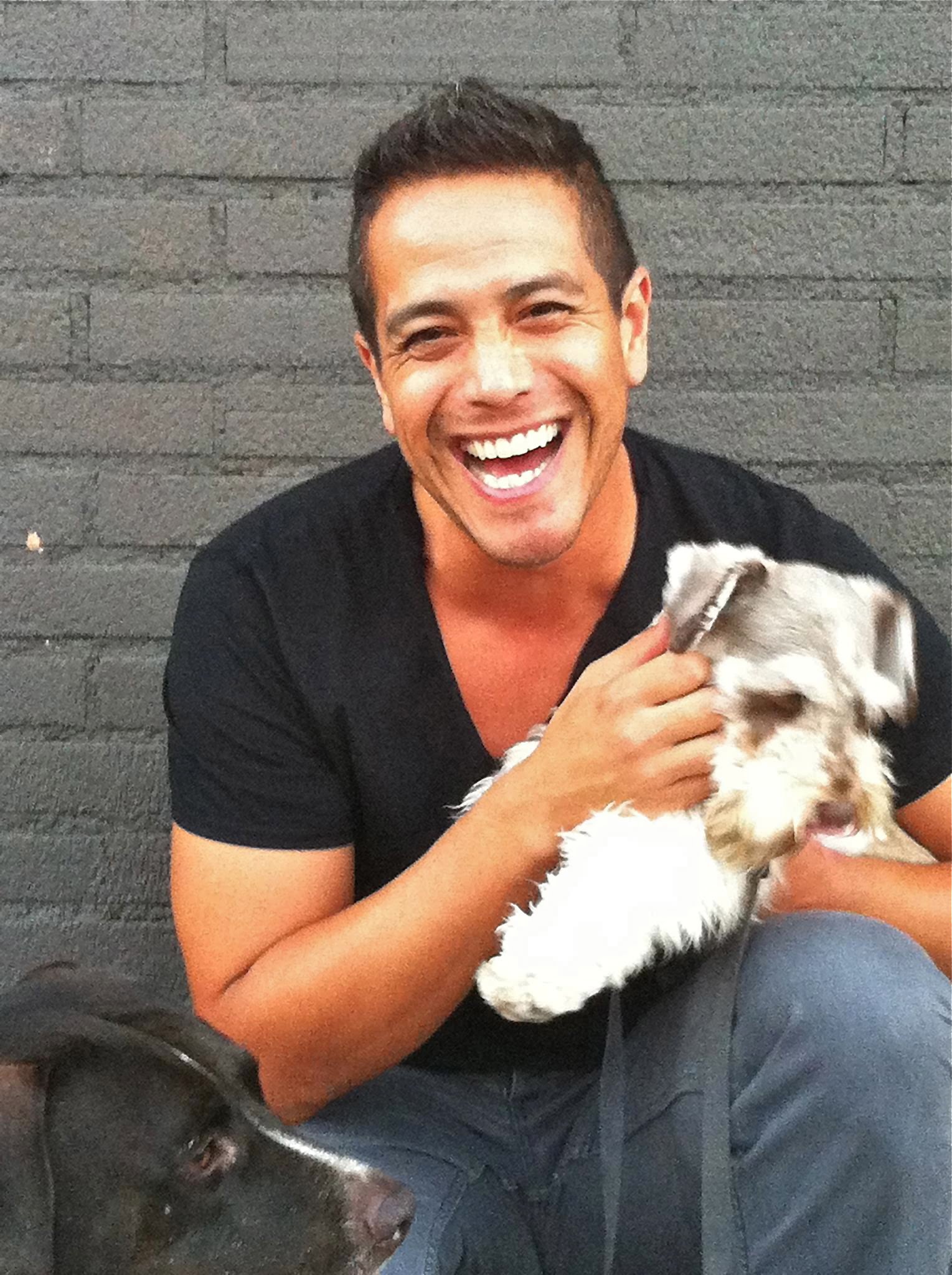 A person is sitting and smiling, holding a small dog in their cozy home. Another dog is visible in the foreground. The background features a charming brick wall.