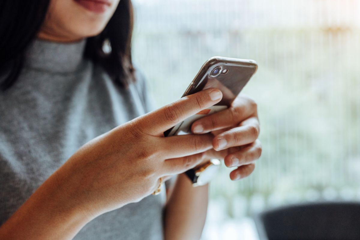 A person holding and using a smartphone with both hands is featured, the focus on the hands and phone while the blurred background hints at a cozy home setting. The individual is dressed in a gray top.